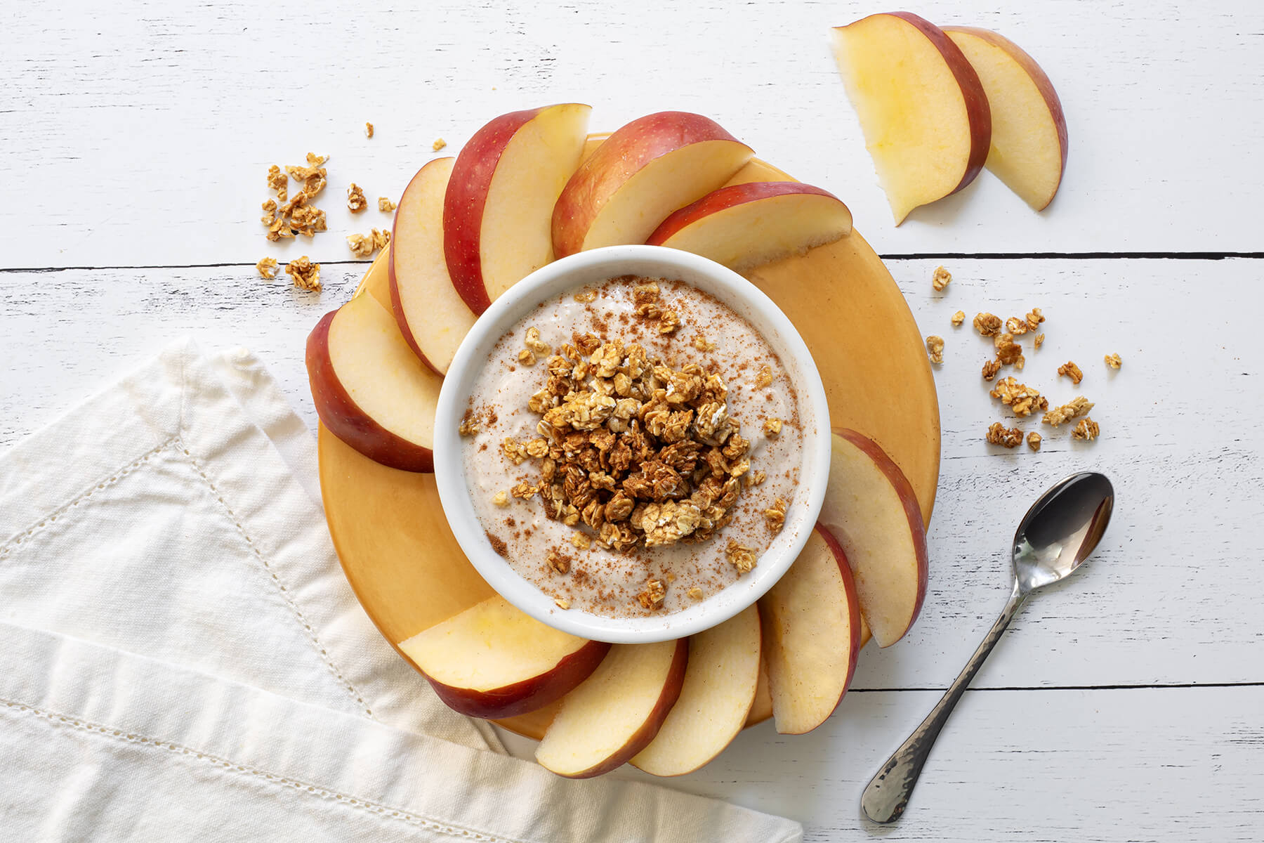 Creamy cinnamon yogurt dip in a white bowl surrounded by fresh apple slices and rice cakes on a wooden serving board.
