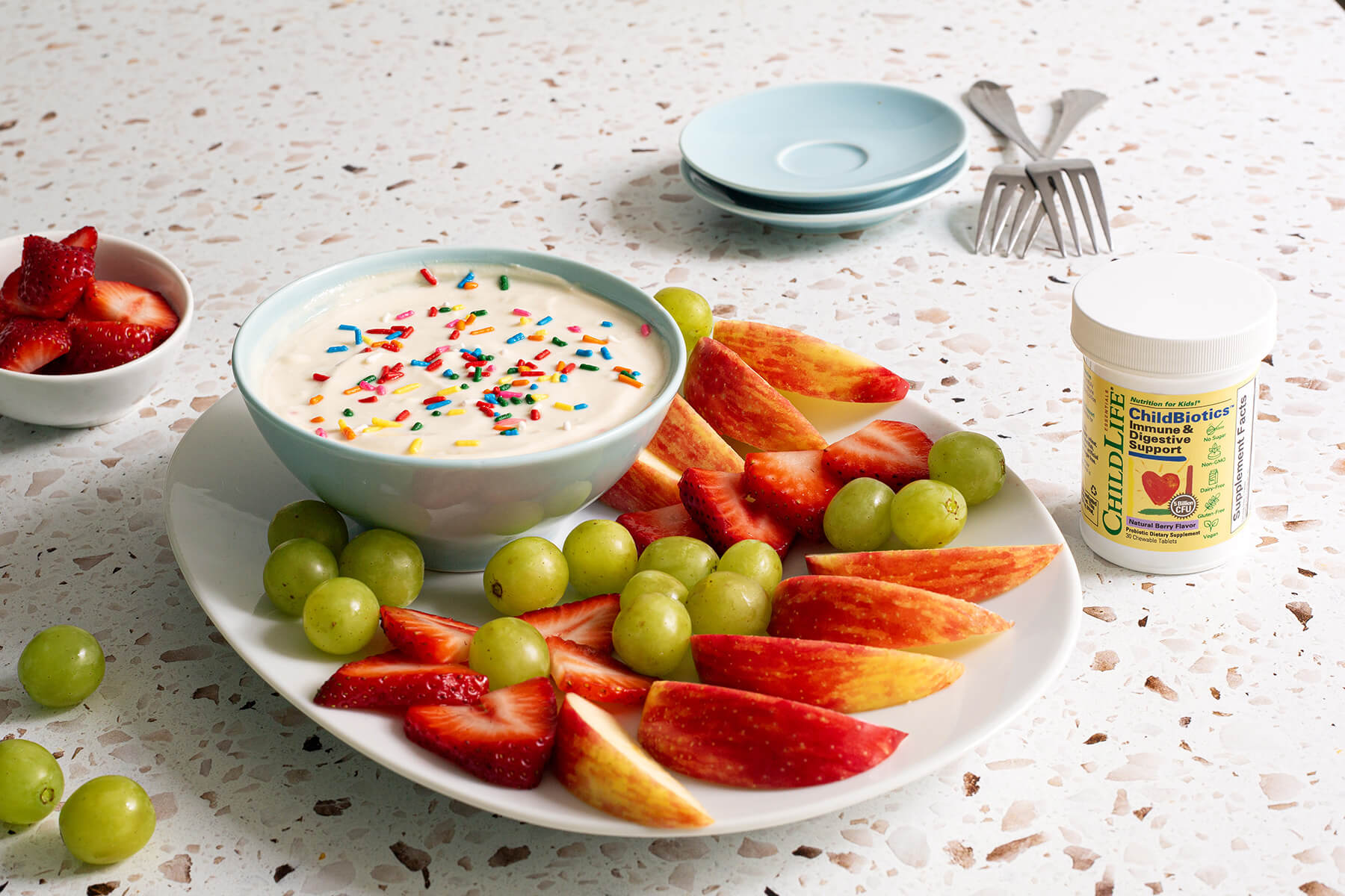 Bowl of creamy yogurt fruit dip topped with rainbow sprinkles, surrounded by fresh strawberries, green grapes, and apple slices on a white platter.