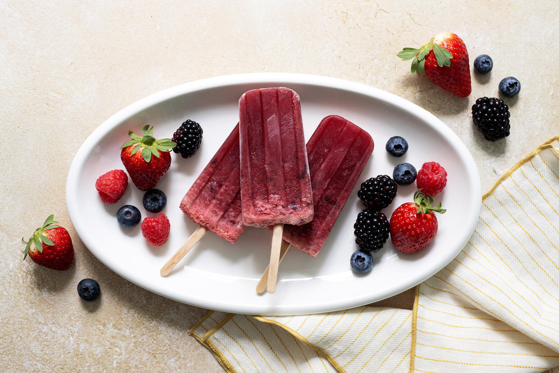 Deep red-purple homemade popsicles made with mixed berries and elderberry, served on a light surface with fresh berries scattered around.