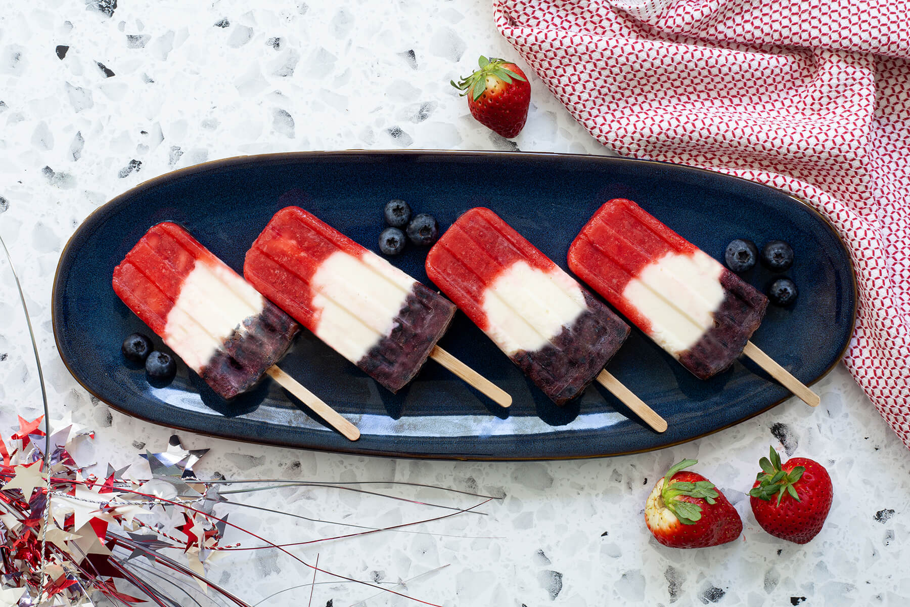 Four homemade red, white, and blue layered popsicles on a navy serving plate with fresh strawberries, blueberries, and a star-spangled napkin.